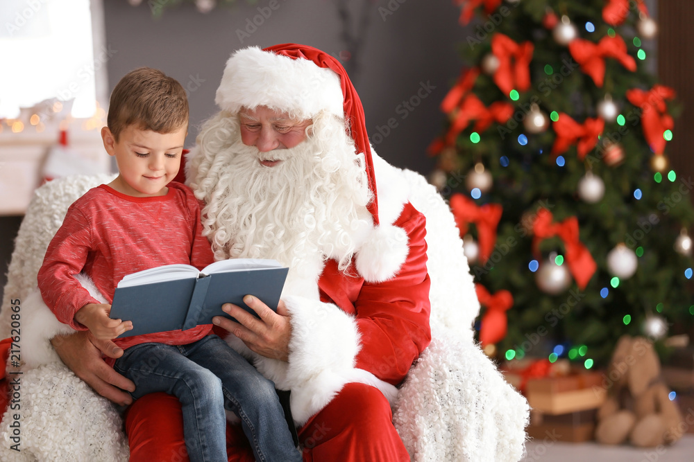 Little boy reading book while sitting on authentic Santa Claus' lap indoors Little boy reading book while sitting on authentic Santa Claus' lap indoors