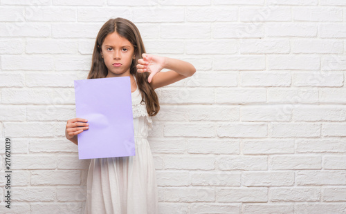 Young hispanic kid over white brick wall holding pink paper sheet with angry face, negative sign showing dislike with thumbs down, rejection concept photo