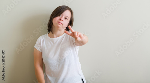 Down syndrome woman standing over wall smiling looking to the camera showing fingers doing victory sign. Number two.
