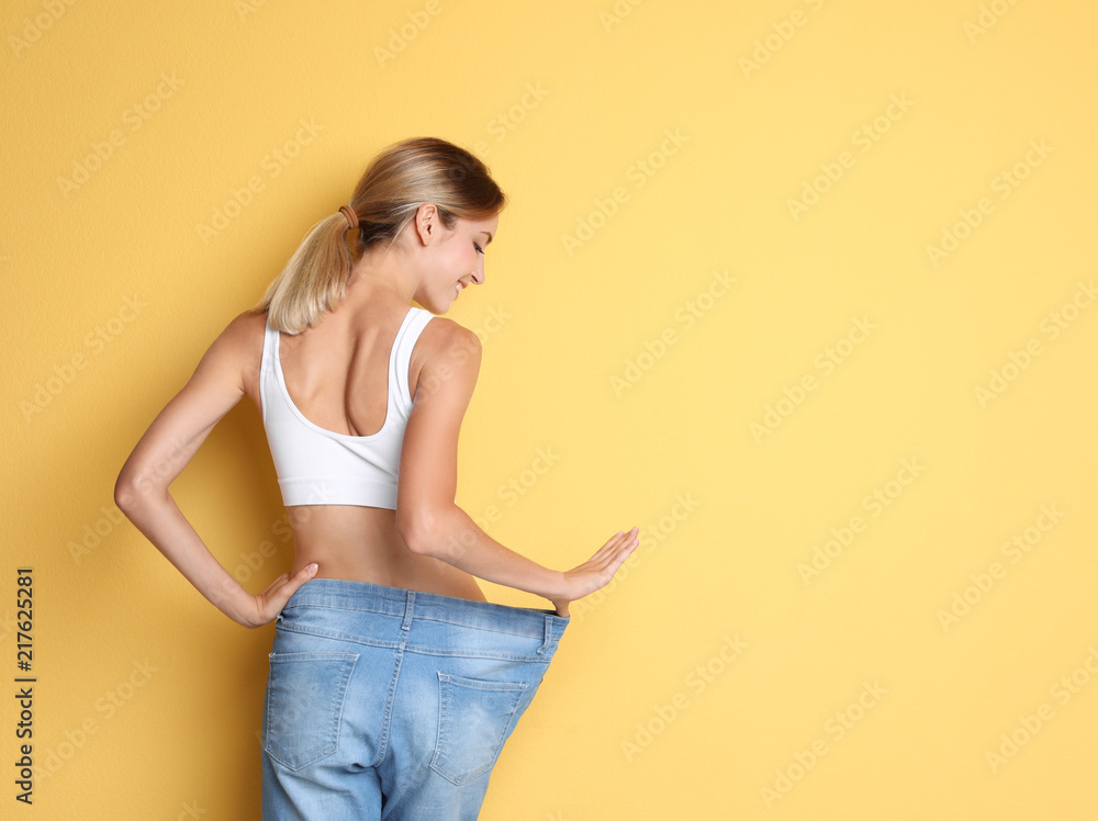 Young slim woman in old big jeans showing her diet results on color background - obrazy, fototapety, plakaty 