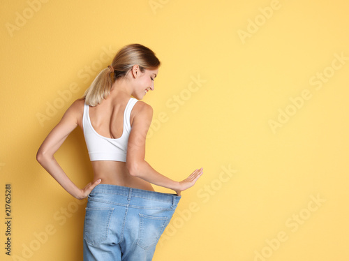 Young slim woman in old big jeans showing her diet results on color background Fototapeta