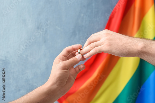 Young man putting wedding ring on his boyfriend's finger near rainbow flag. Gay marriage photo