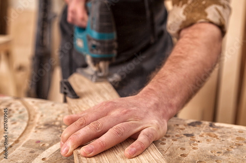 Close up of experienced carpenter in work clothes and small buiness owner carpenter saw and processes the edges of a wooden bar with a jig saw in a light workshop