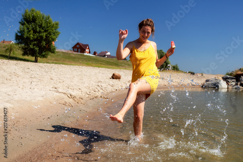 Εκτύπωση καμβά A cheerful dark-haired woman in summer dress eat watermelon, walks along the beach, kicks waves, on a summer day