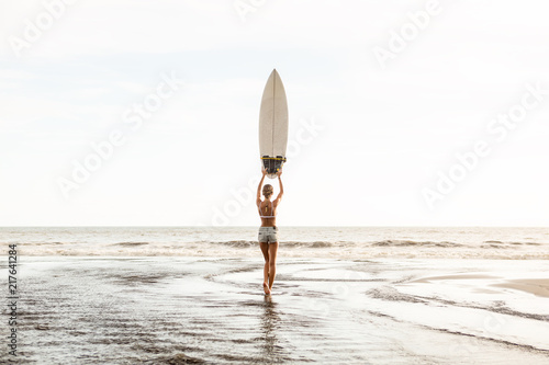 Young surfer girl in sexy white bikini and short denim shorts stay in river and pose with surf short board. Modern family lifestyle, people water sport adventure camp, extreme swim on summer vacation photo