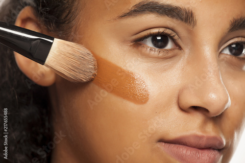 portrait of a young dark-skinned woman applying liquid makeup base with brush on her face on a white background