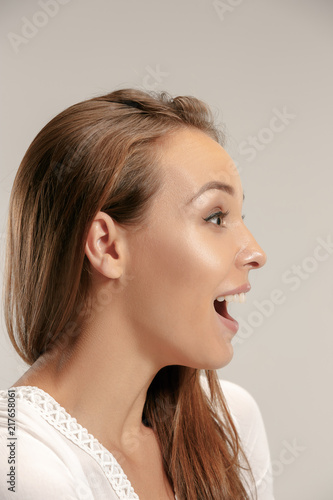 Wow. Beautiful female face isolated on gray studio backgroud. Young emotional surprised woman standing with open mouth. Human emotions, facial expression concept. Profile
