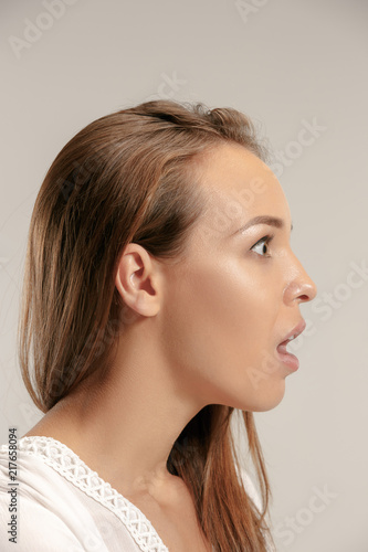 Wow. Beautiful female face isolated on gray studio backgroud. Young emotional surprised woman standing with open mouth. Human emotions, facial expression concept. Profile