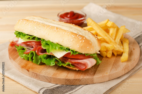 Fresh submarine sandwich with ham, cheese, bacon, tomatoes and  lettuce on wooden cutting board with french fries and sauce