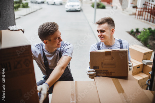 Two young handsome smiling movers wearing uniforms are unloading the van full of boxes. House move, mover service.