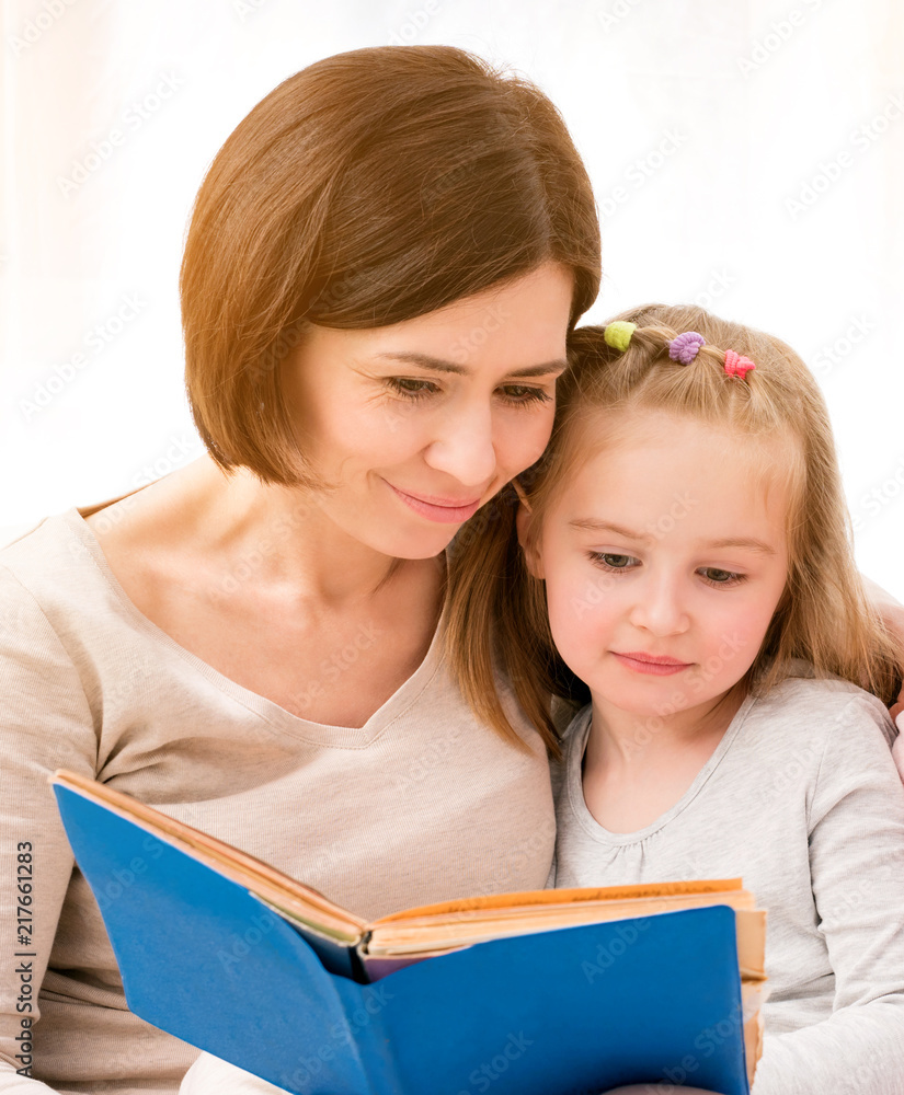 Mother and little daughter reading book sitting on sofa in light living room. Happy family time together Mother and little daughter reading book sitting on sofa in light living room. Happy family time together