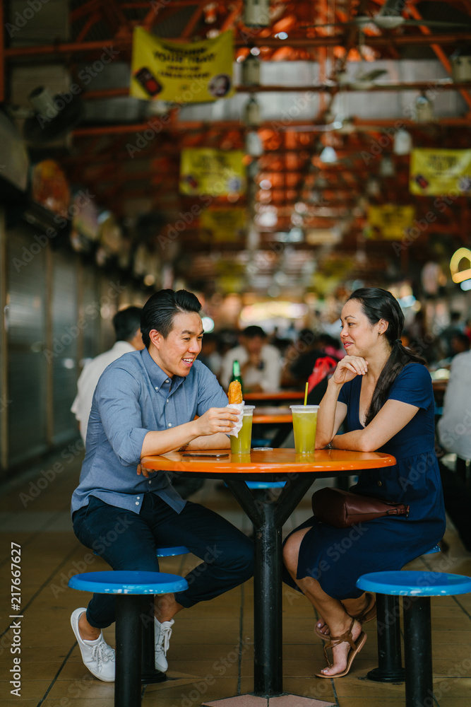 Image of a Chinese Asian young couple enjoying a snack together at a hawker center in Singapore, Asia. They are enjoying their sugarcane juice and fried bananas while having a fun conversation. 