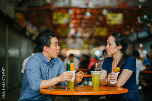 Image of  a good looking Chinese Asian young couple enjoying a snack at a hawker center Singapore  Asia. They are smiling as they converse and enjoy their sugarcane juice and fried bananas. 