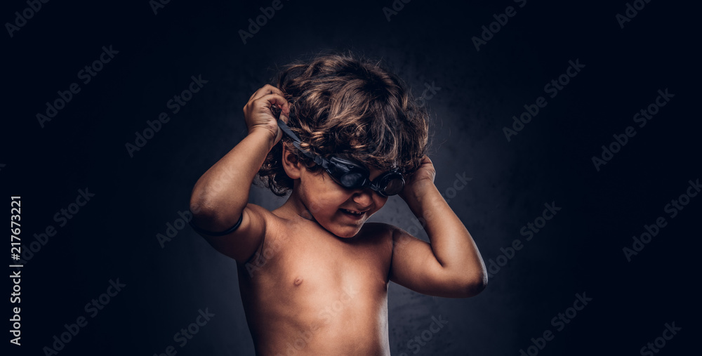 Cute little shirtless boy puts on swimming goggles posing in a studio. Isolated on the dark textured background.
