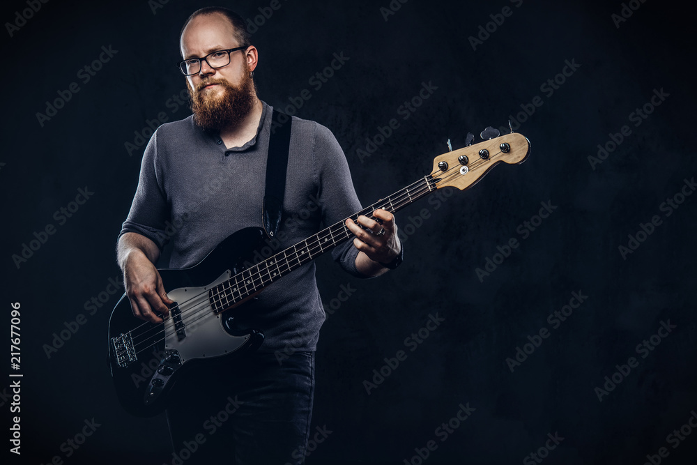 Obraz premium Redhead bearded male musician wearing glasses dressed in a gray t-shirt playing on electric guitar. Isolated on dark textured background.