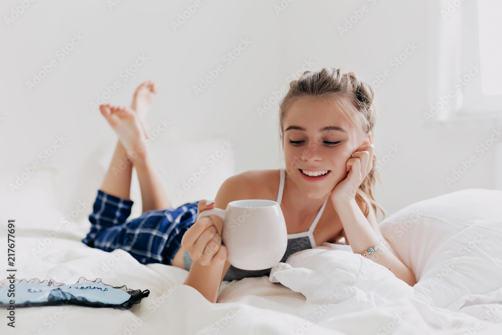 Young attractive happy woman with a cup of coffee lying in the bed by the window, closed eyes and enjoys of her morning. Top view from above. Lazy day off concept Young attractive happy woman with a cup of coffee lying in the bed by the window, closed eyes and enjoys of her morning. Top view from above. Lazy day off concept
