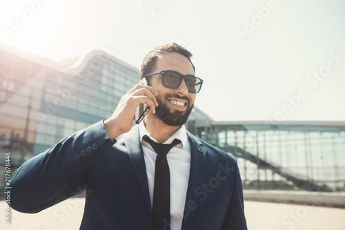 Bearded smiling businessman wears blue suit and sunglasses talking via smartphone before high modern building