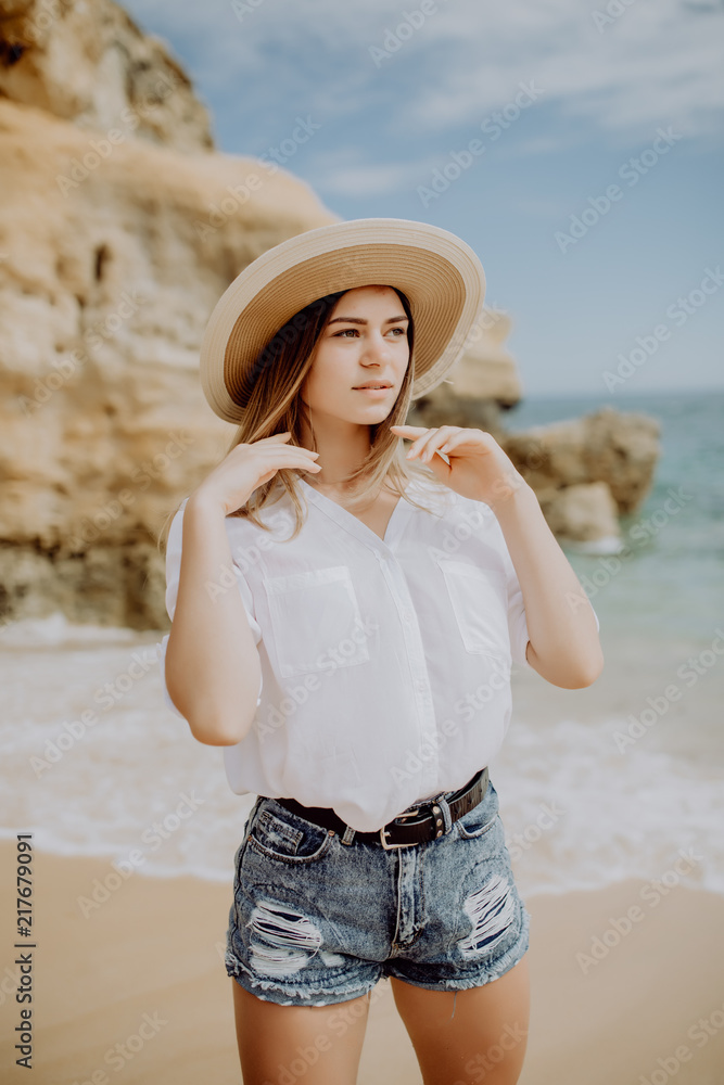 Young beautiful woman wearing straw hat posing standing on beach near waves and rock and looking away at the sea