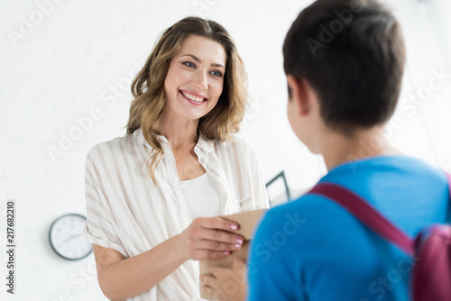 smiling mother giving paper package with meal to son with backpack at home  back to school concept