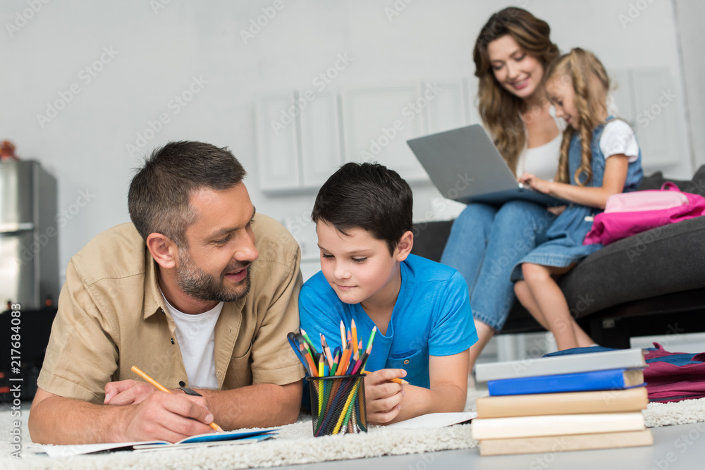 selective focus of man helping son with homework while mother and daughter using laptop on sofa together at home selective focus of man helping son with homework while mother and daughter using laptop on sofa together at home