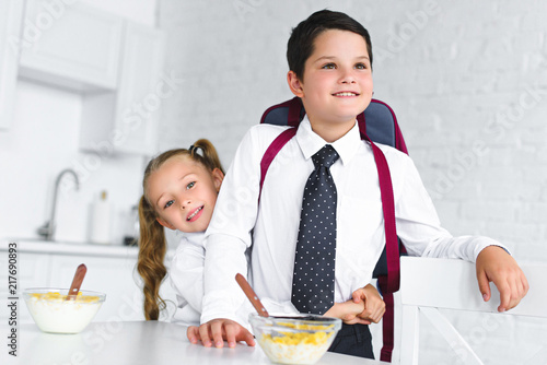 little sister hugging brother in school uniform with backpack at table with breakfast in kitchen at home  back to school concept