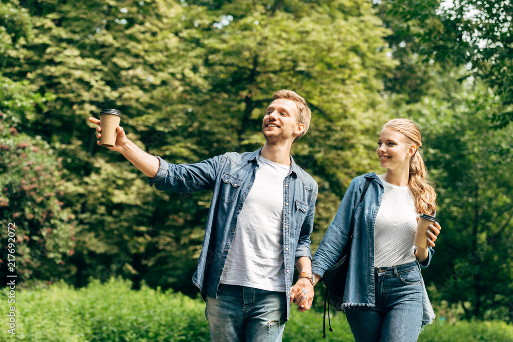 happy young couple with paper cups of coffee walking by park together and pointing somewhere