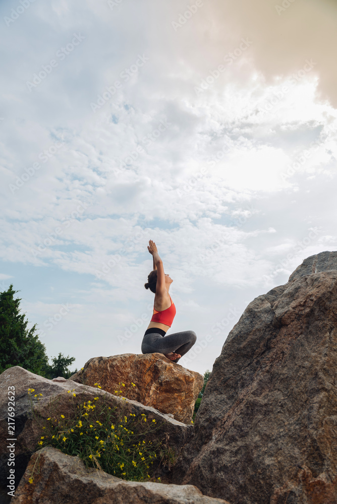 Flexible and slim. Flexible and slim woman lifting her hands while doing yoga sitting on rock around nature