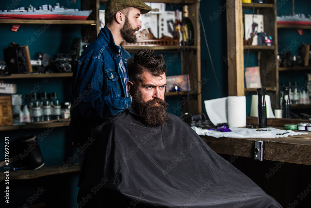 Hipster client getting haircut. Client with beard ready for trimming or grooming. Man with beard covered with black cape waiting while barber changing clipper grade. Haircut process concept Hipster client getting haircut. Client with beard ready for trimming or grooming. Man with beard covered with black cape waiting while barber changing clipper grade. Haircut process concept