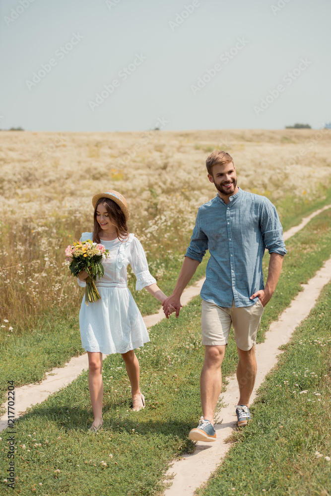 woman in white dress with bouquet of wild flowers walking together with boyfriend in field woman in white dress with bouquet of wild flowers walking together with boyfriend in field