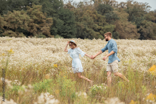 side view of young lovers holding hands while walking together in field with wild flowers