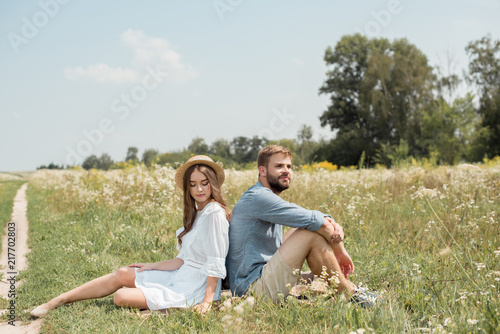 young couple resting back to back on blanket in field with wild flowers on summer day