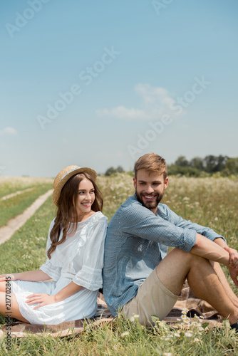 young couple resting back to back on blanket in field with wild flowers on summer day