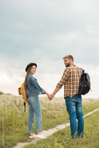 smiling couple with backpacks holding hands in summer field with wild flowers