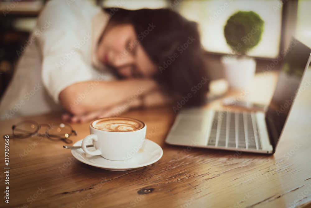 Close up of full of coffee in coffee cup with business woman tired from working and laptop computer background. Relax Sleeping and Healthy concept. Business people and overtime workload theme Close up of full of coffee in coffee cup with business woman tired from working and laptop computer background. Relax Sleeping and Healthy concept. Business people and overtime workload theme