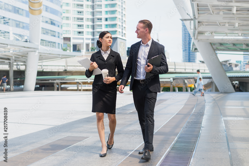 Asian business woman holding document file and plastic mug on hand talk about business future with Caucasian businessman. they are walk together in the city. Asian business woman holding document file and plastic mug on hand talk about business future with Caucasian businessman. they are walk together in the city.