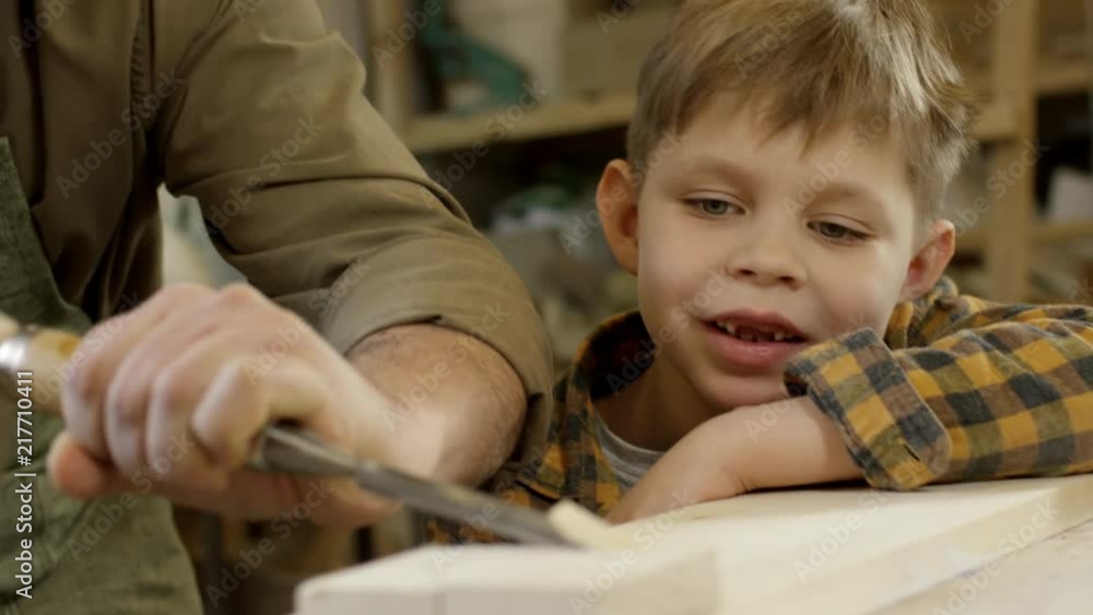 Tilt down with rack focus of curious little boy looking at cheerful male carpenter in apron using chisel and mallet to carve wood Tilt down with rack focus of curious little boy looking at cheerful male carpenter in apron using chisel and mallet to carve wood