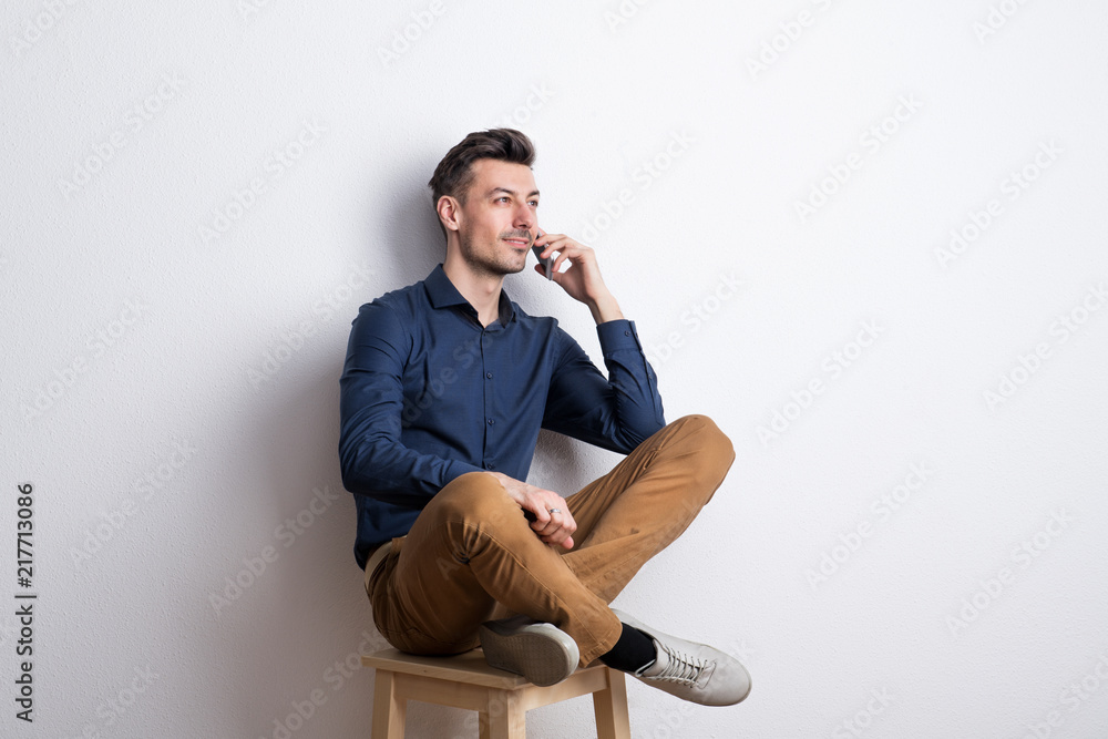 Young man with smartphone in a studio sitting on a stool, making a phone call. Young man with smartphone in a studio sitting on a stool, making a phone call.