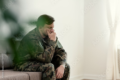 Sad soldier in uniform covering his mouth while sitting on a sofa © Photographee.eu