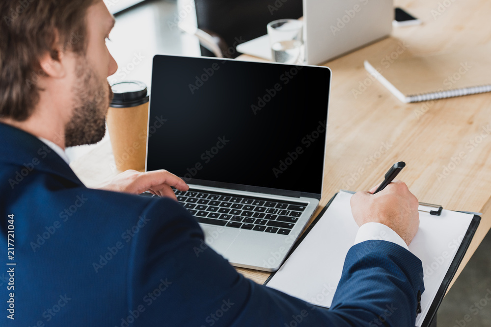 cropped shot of businessman taking notes on clipboard and using laptop with blank screen at workplace cropped shot of businessman taking notes on clipboard and using laptop with blank screen at workplace