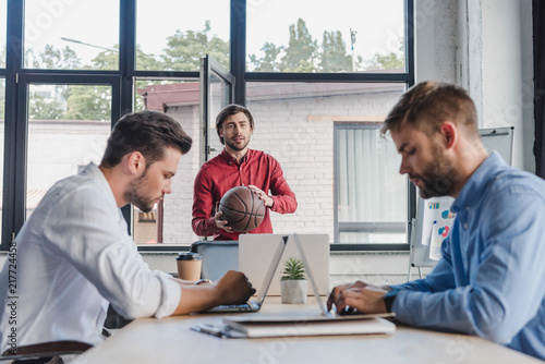 Fototapeta Naklejka Na Ścianę i Meble -  young businessman holding basketball ball while colleagues using laptops in office