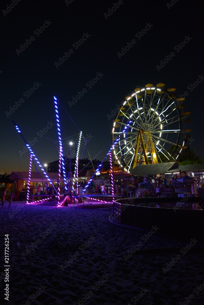 Night entertainment and relaxation on the beach of the Black Sea with attractions, white Ferris wheel, trampoline with a light, blue, yellow color and a children's pool