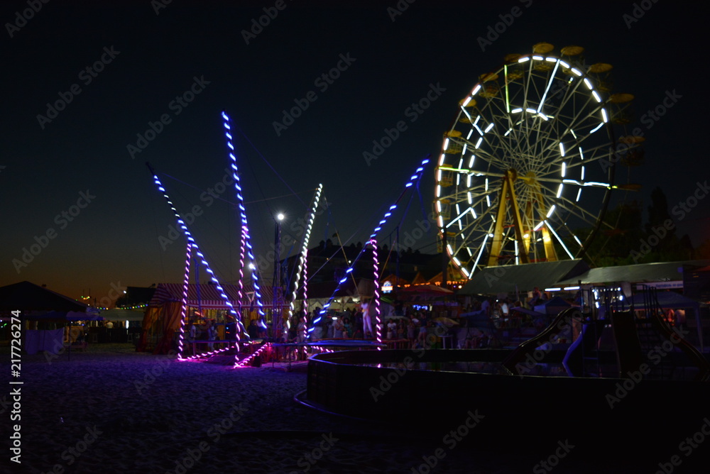 Night entertainment and relaxation on the beach of the Black Sea with attractions, white Ferris wheel, trampoline with a light, blue, yellow color and a children's pool Night entertainment and relaxation on the beach of the Black Sea with attractions, white Ferris wheel, trampoline with a light, blue, yellow color and a children's pool