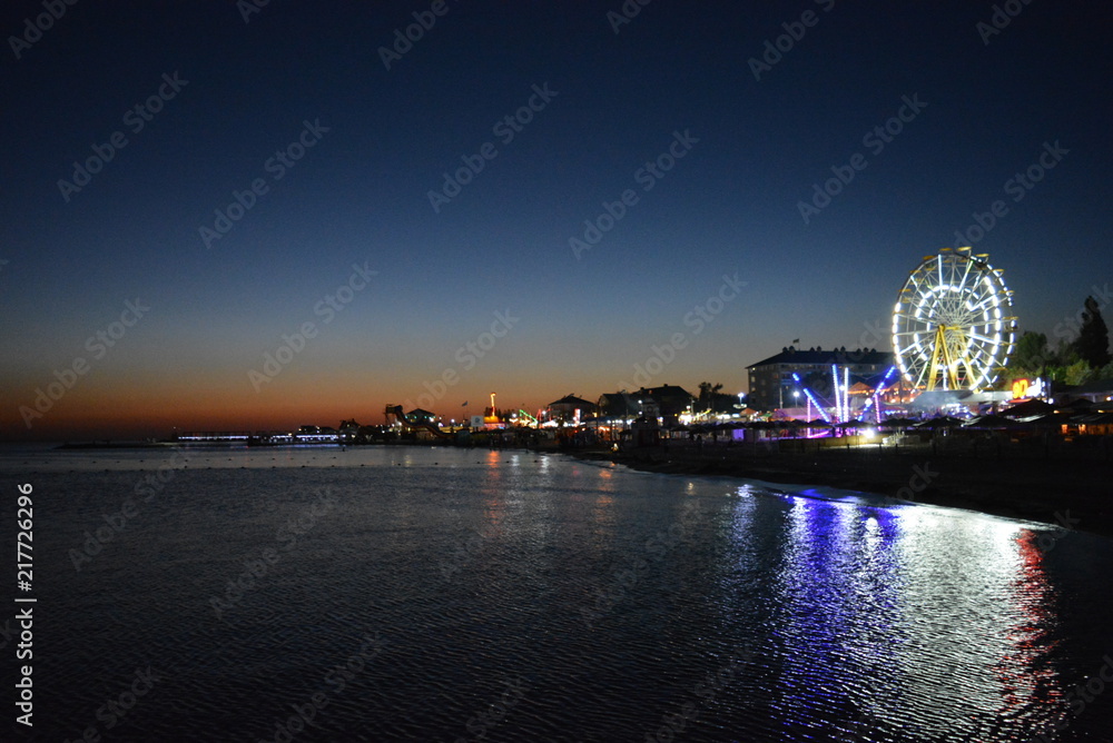 Beautiful sea water with reflection and the embankment of the Black Sea, houses with a Ferris wheel and bright lanterns at sunset at sunset