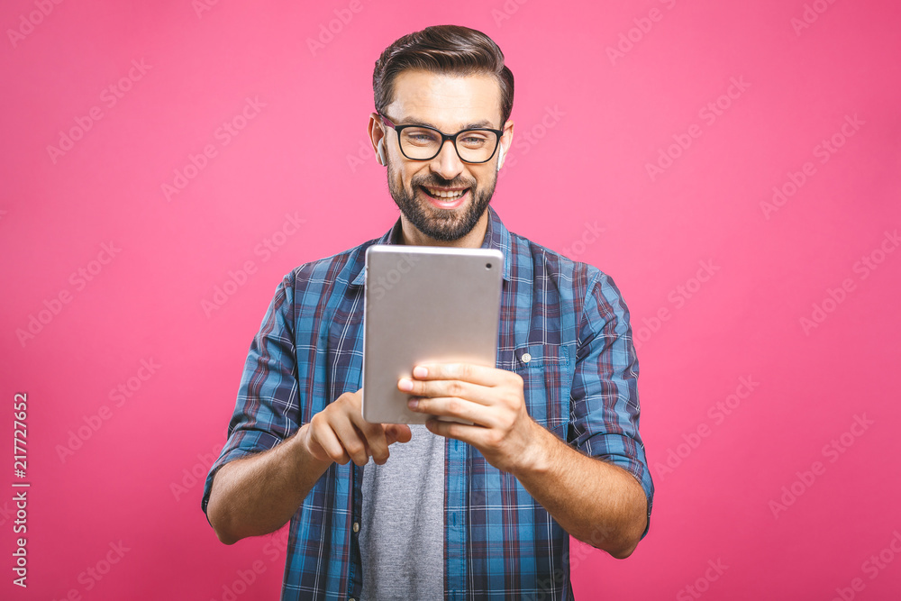 Happy young man in plaid shirt standing and using tablet over pink background Happy young man in plaid shirt standing and using tablet over pink background