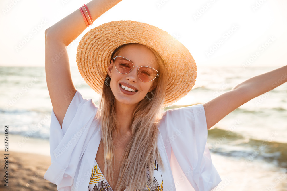 Image of beautiful blonde woman 20s in summer straw hat and sunglasses smiling, while resting at seaside