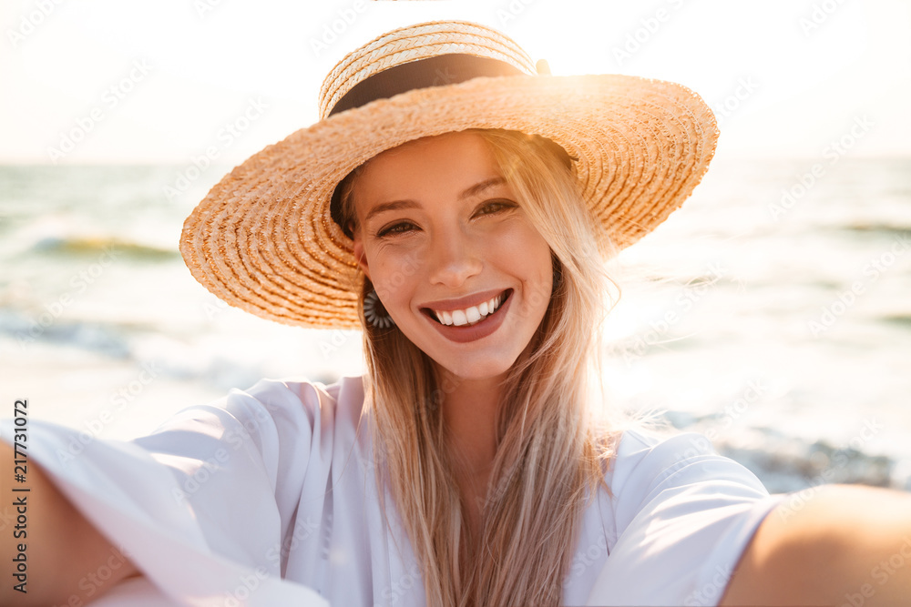 Portrait of positive blonde woman 20s in summer straw hat smiling, and taking selfie while walking at sea coast