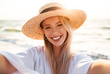 Portrait of positive blonde woman 20s in summer straw hat smiling, and taking selfie while walking at sea coast