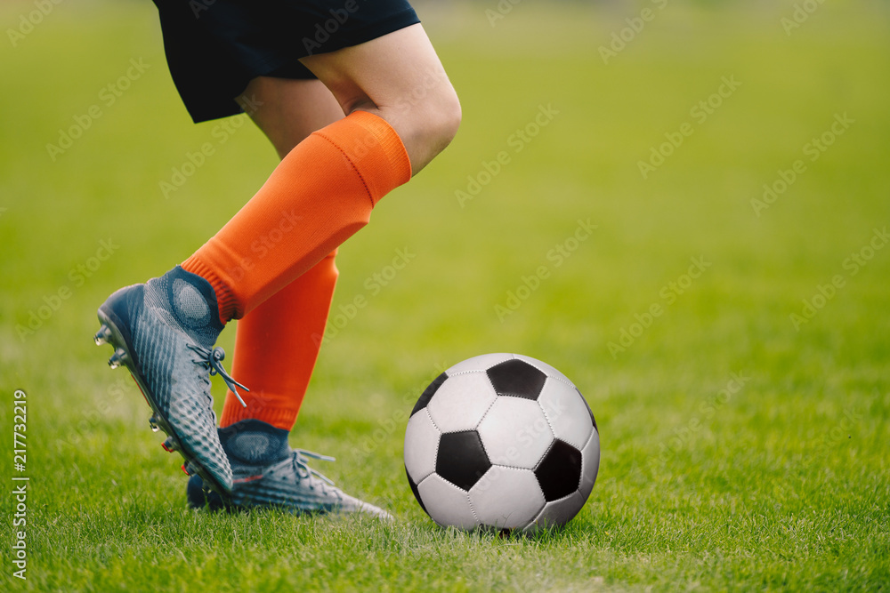 A young man kicking a football outdoors. Young football player training on a grass field. Boy in a sportswear. Soccer player wearing orange socks and soccer boots. Football horizontal background