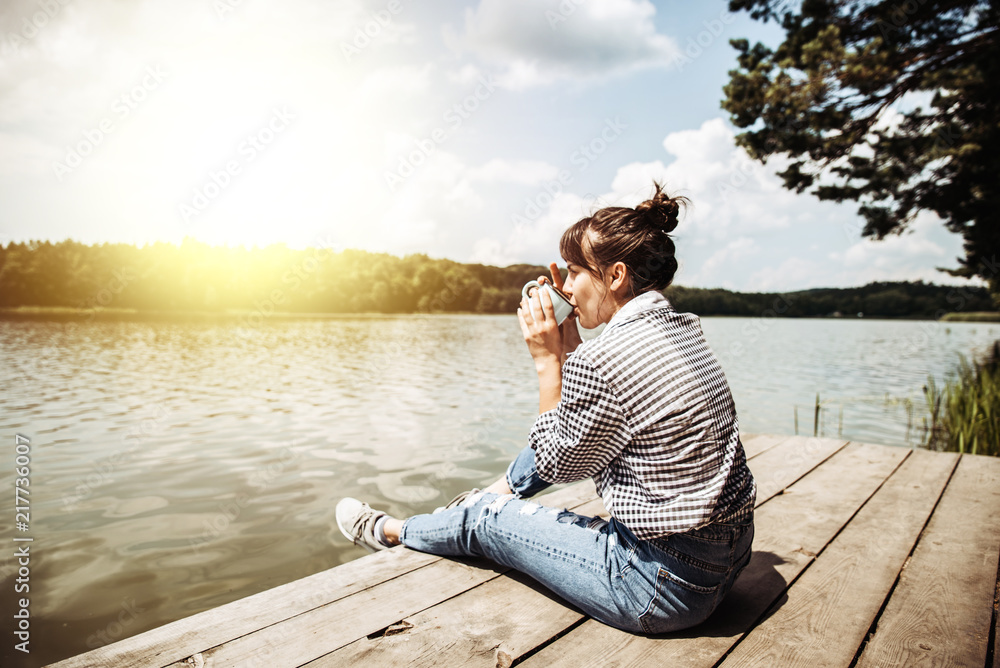 young adult woman sitting on wooden dock drinking coffee and loo