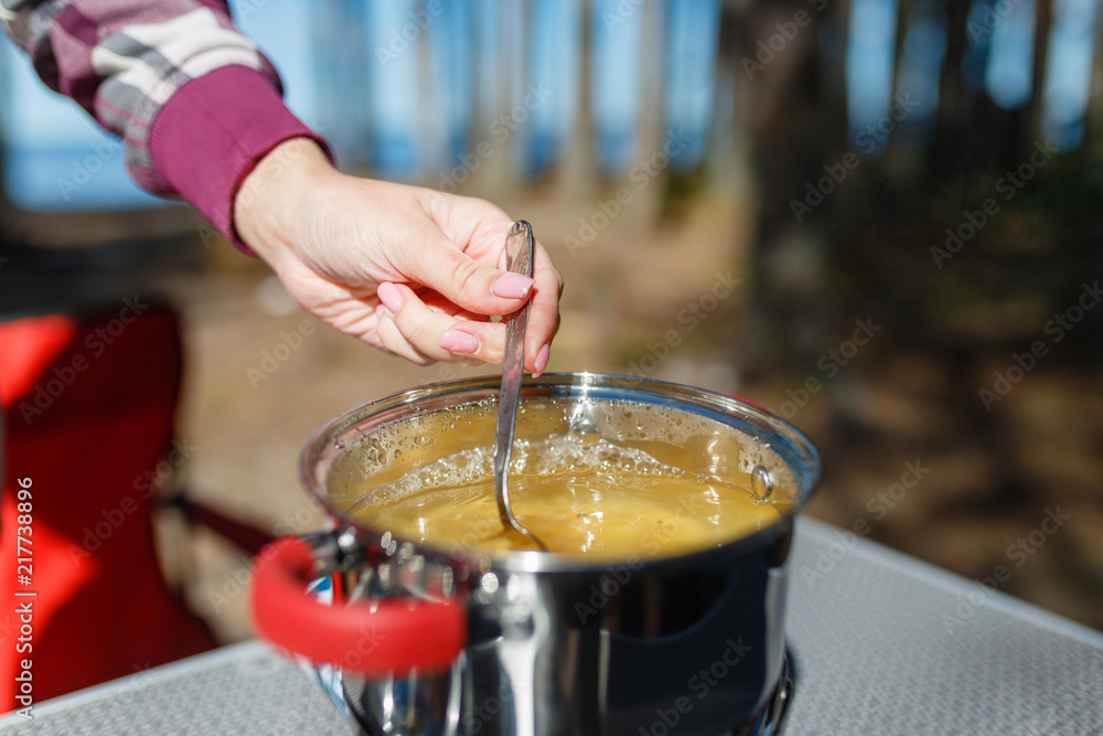 Girl traveler prepares food on portable gas stove, on a folding table on the background of camping in forest. Women's hands interfere with a spoon food in a pot on a gas burner (camping stove).